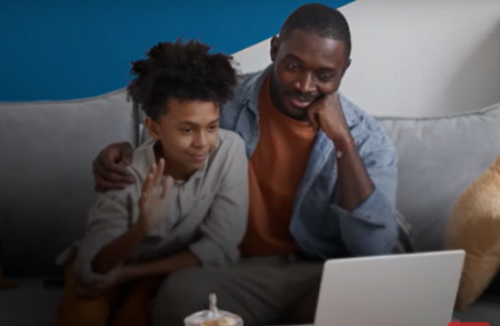 Un padre y su hijo sentados en un sillón y saludando a un profesional de la salud que aparece en la pantalla de una computadora.