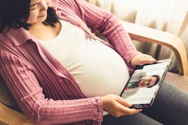 Primer plano de una mujer embarazada sentada en un sofá, sujetando una tableta sobre el regazo. En la pantalla de la tableta se puede ver a un profesional de la salud.