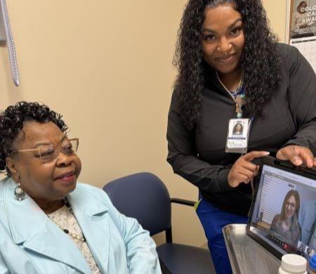 Woman sitting in a chair while a nurse holds a laptop with live video of physician.